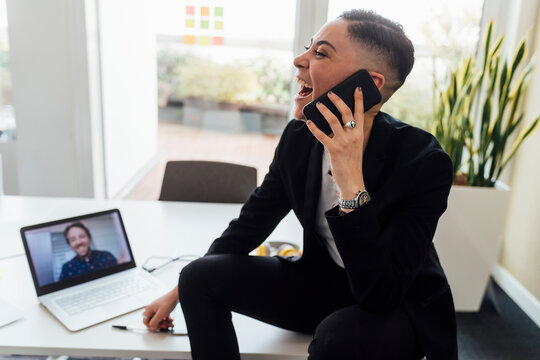 Happy businesswoman talking on mobile phone during video call through laptop at office - Powered by Adobe