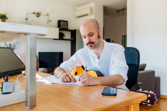 Male music composer with guitar writing musical notes at table in living room
