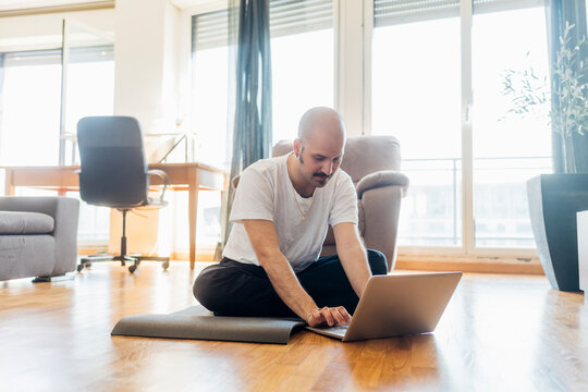 Young man using laptop while sitting on exercise mat at home