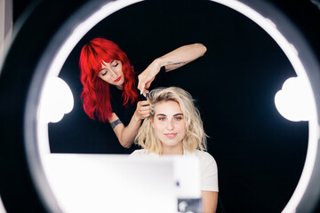 Female stylist doing hairstyle of beautiful model while recording through mobile phone in studio