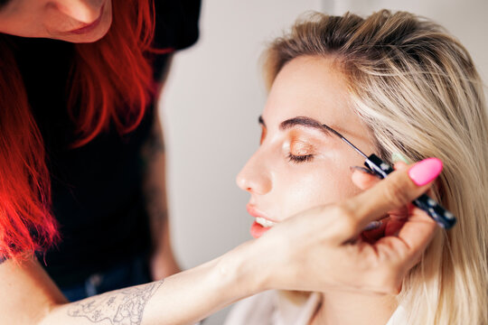 Young female make-up artist working on eyebrows of model with eyes closed at studio