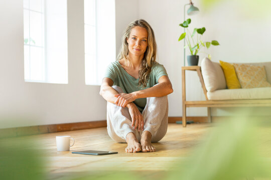 Confident woman in loungewear sitting on floor by digital tablet and coffee mug at home