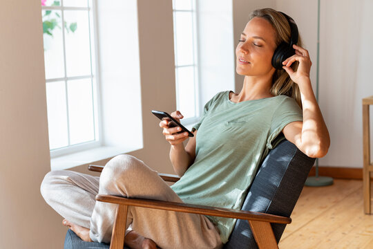 Woman with eyes closed listening music and holding mobile phone while resting at home