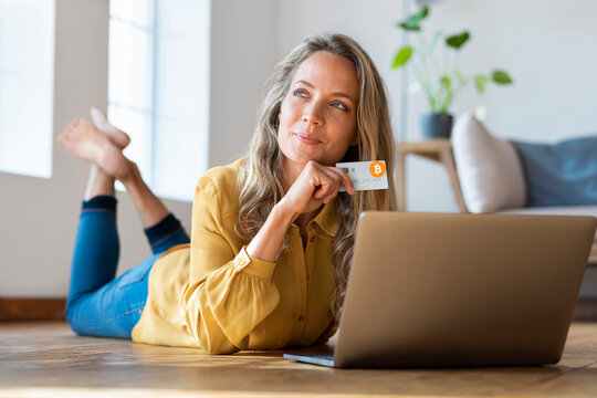 Contemplating woman with cryptocurrency credit card looking away while lying down in front of laptop at home