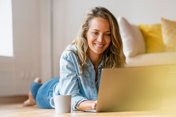 Smiling woman lying on hardwood floor while using laptop at home