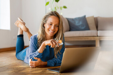 Smiling beautiful woman holding credit card while lying down on floor in front of laptop at home