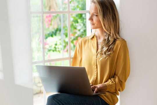 Contemplating woman looking away while using laptop by window at home