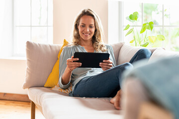 Contented woman looking at digital tablet while sitting on sofa in living room