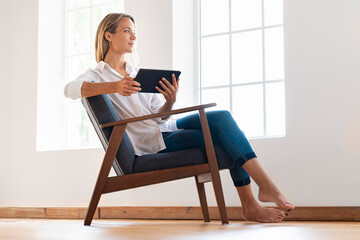Contemplating woman holding digital tablet while sitting on armchair at home