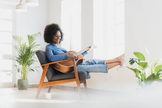 Smiling young woman playing guitar while sitting on armchair at home