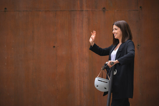 Young businesswoman waving hand while standing with electric push scooter by wall
