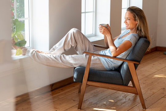 Happy woman holding coffee mug while sitting at window in living room