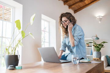 Smiling female professional talking on smart phone while working on laptop at desk
