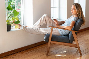 Contemplating woman with mobile phone sitting on armchair at home