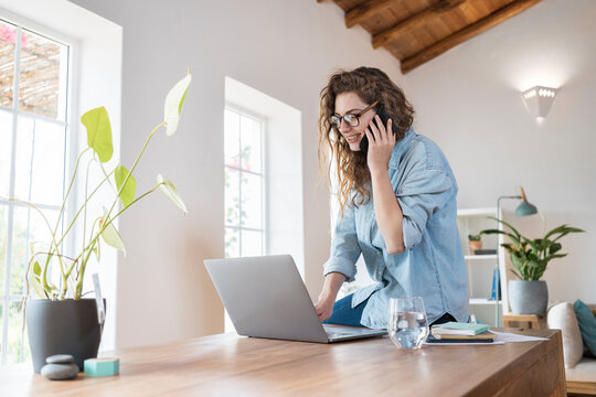 Smiling female professional talking on smart phone while working on laptop at desk