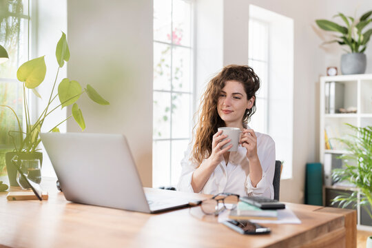 Beautiful female freelance worker having coffee while looking at laptop in home office