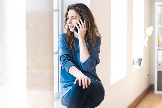 Young woman looking through window while talking on mobile phone at home