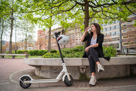 Young businesswoman drinking coffee while sitting by electric push scooter