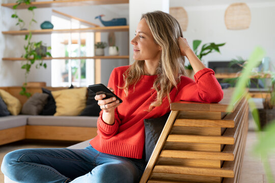Happy woman sitting on couch with mobile phone while looking away