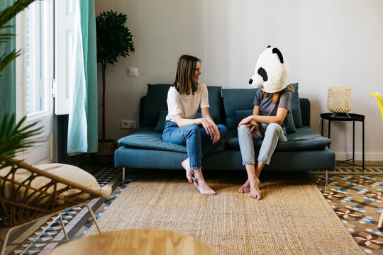 Smiling woman looking at girlfriend wearing panda mask in living room