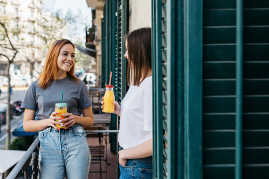 Smiling woman enjoying juice with girlfriend while standing at balcony