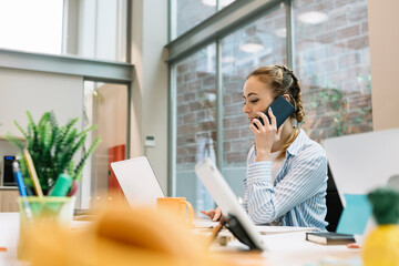 Female professional with laptop talking on mobile phone at office