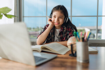 Asian teen girl wearing headphones learning language online, using laptop, looking at screen, doing school tasks at home, writing notes, listening to lecture or music, distance education