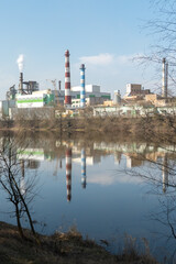 Fototapeta na wymiar A large factory is located on the river bank. Toxic white smoke escapes from the factory's chimneys. The risk of an environmental disaster. Reflection in the water of the river of the plant