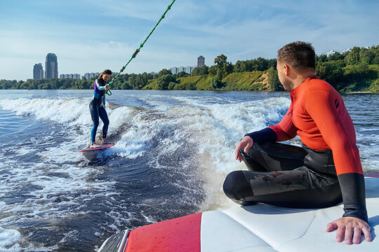 Man sitting at edge of moving boat looking at female wakeboarder surfing behind