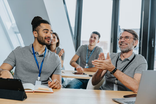 Male trainee looking at smiling colleague clapping while sitting on bench in training class