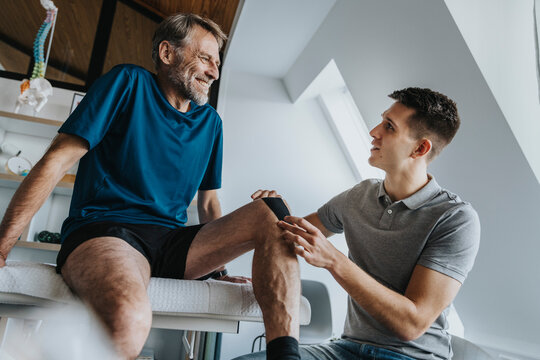 Male physical therapist sticking kinesio tape on smiling patient's knee in practice