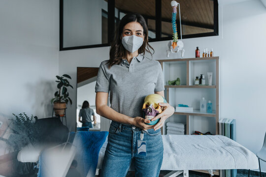 Female physiotherapist wearing protective face mask holding skull model in practice