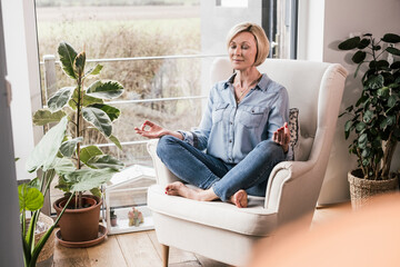 Mature woman meditating while sitting on armchair in living room