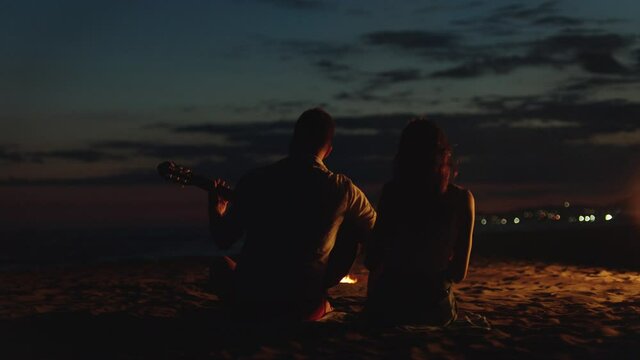 Back View Of Couple Sitting Around Campfire, Playing Guitar, Singing And Leaning To Rhythm. Summer Evening At Seaside.