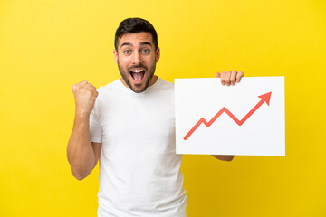 Young handsome caucasian man isolated on yellow background holding a sign with a growing statistics arrow symbol and celebrating a victory