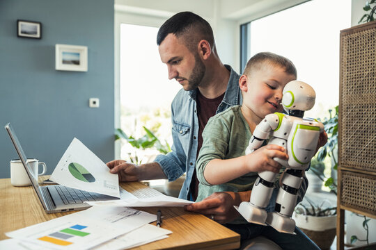 Boy playing with robot sitting while businessman analyzing graph at home