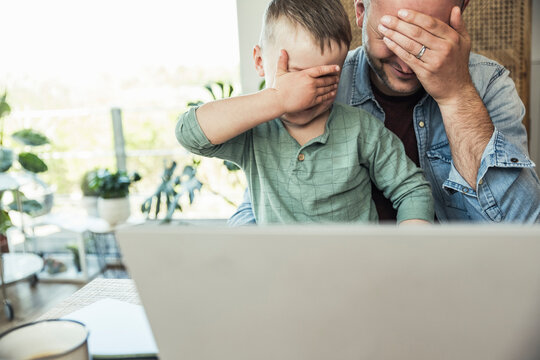 Father and son covering eyes with hands while sitting with laptop at home