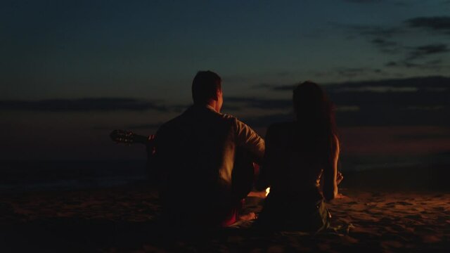 Low light scene of couple sitting around campfire from behind. Playing guitar, singing and leaning to rhythm. Summer evening at seaside.