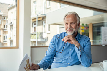 Smiling businessman holding eyeglasses and documents while sitting at desk in office