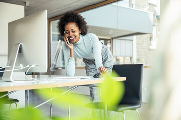 Smiling businesswoman leaning on desk while talking on mobile phone at office