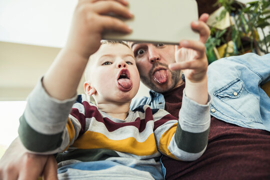 Father sticking out tongue with son talking selfie through smart phone at home