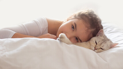 Obraz premium Wide banner. A beautiful little girl in white clothes, lies on a clean, white bed with a white British cat.