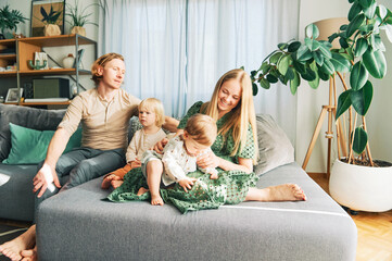 Happy young family of four relaxing on a couch, couple playing with baby girl and toddler boy