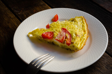 A serving of rustic omelet in a white plate, bread, kitchen napkin on a wooden table.