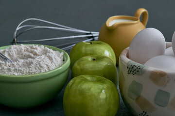 Green apples, milk, flour, white eggs in a colorful bowl on a textured gray background