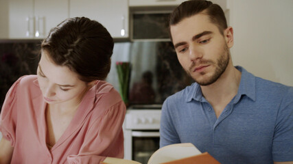 A young man is reading a book while his wife is writing something down on paper