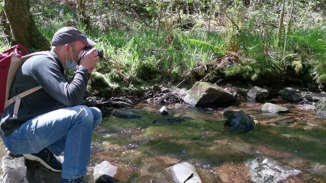 Man in cap crouched down taking pictures with mask in a river. People in nature.