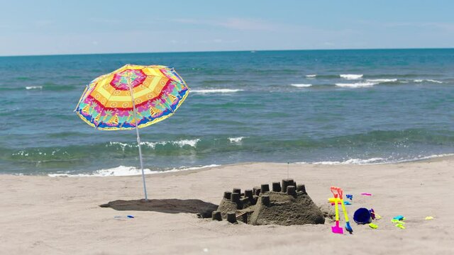Sandcastle on beach, child toys and colourful parasol around. Sea waves splashing in background. Sunny summer day at seaside.