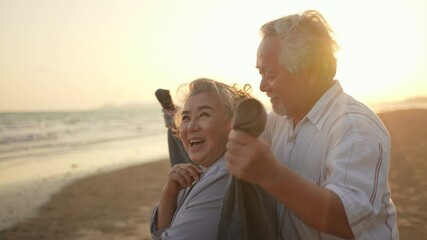 Happy Asian family senior husband cover his wife with a blanket and hugging together on the beach at summer sunset. Healthy elderly retirement couple relax and enjoy romantic outdoor vacation together - Powered by Adobe