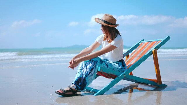 Beautiful Asian woman tourist resting on sun bed by the sea. Happy female in casual clothing sit on beach chair relax and enjoy outdoor lifestyle at tropical beach in summer holiday travel vacation